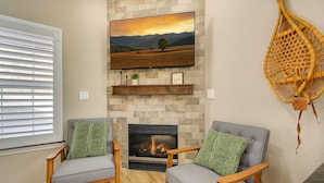 Een woonkamer met een open haard, twee fauteuils, een televisie boven de open haard en een decoratieve mand aan de muur.