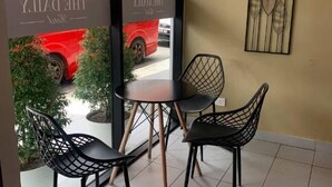 Een café-interieur met een tafel en twee stoelen, een raam met uitzicht op een rode auto en een kunstwerk aan de muur.