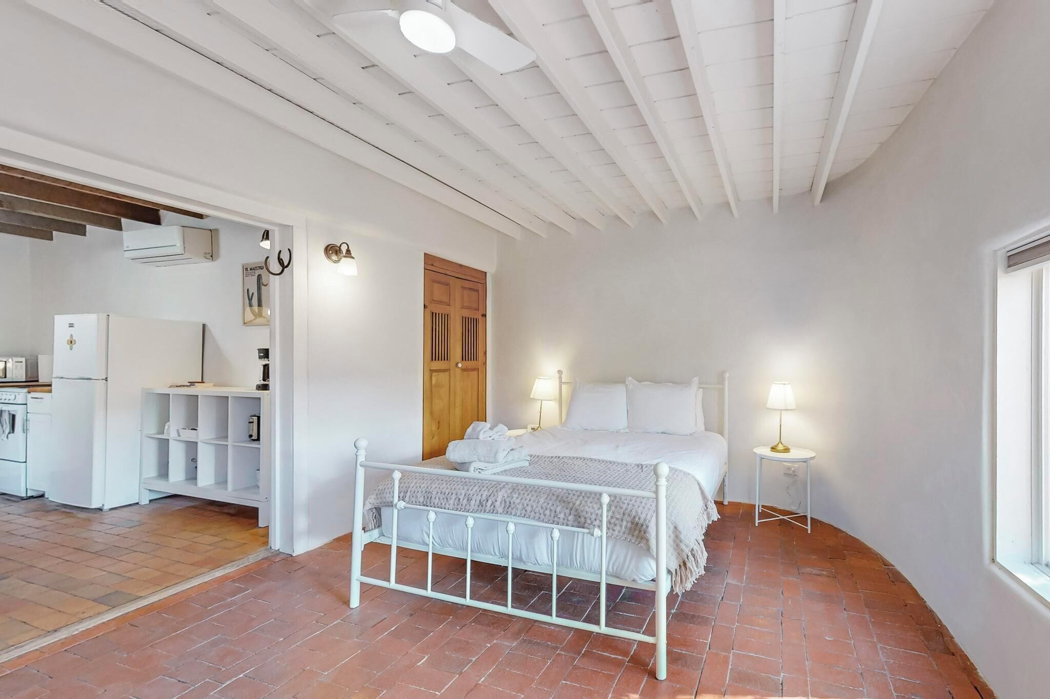 Quarto com cama branca, duas luminárias de cabeceira, porta de madeira e uma pequena mesa branca com luminária.