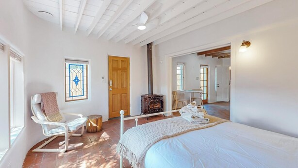 A room with a bed, a white rocking chair, a wooden door, a stove, and a small table with chairs.