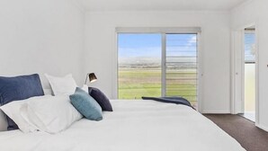 A bedroom with a large bed, a nightstand, and a view of a green landscape through a sliding glass door. - Sea View at Silver Sands - C21 SouthCoast Holidays (Aldinga Beach)