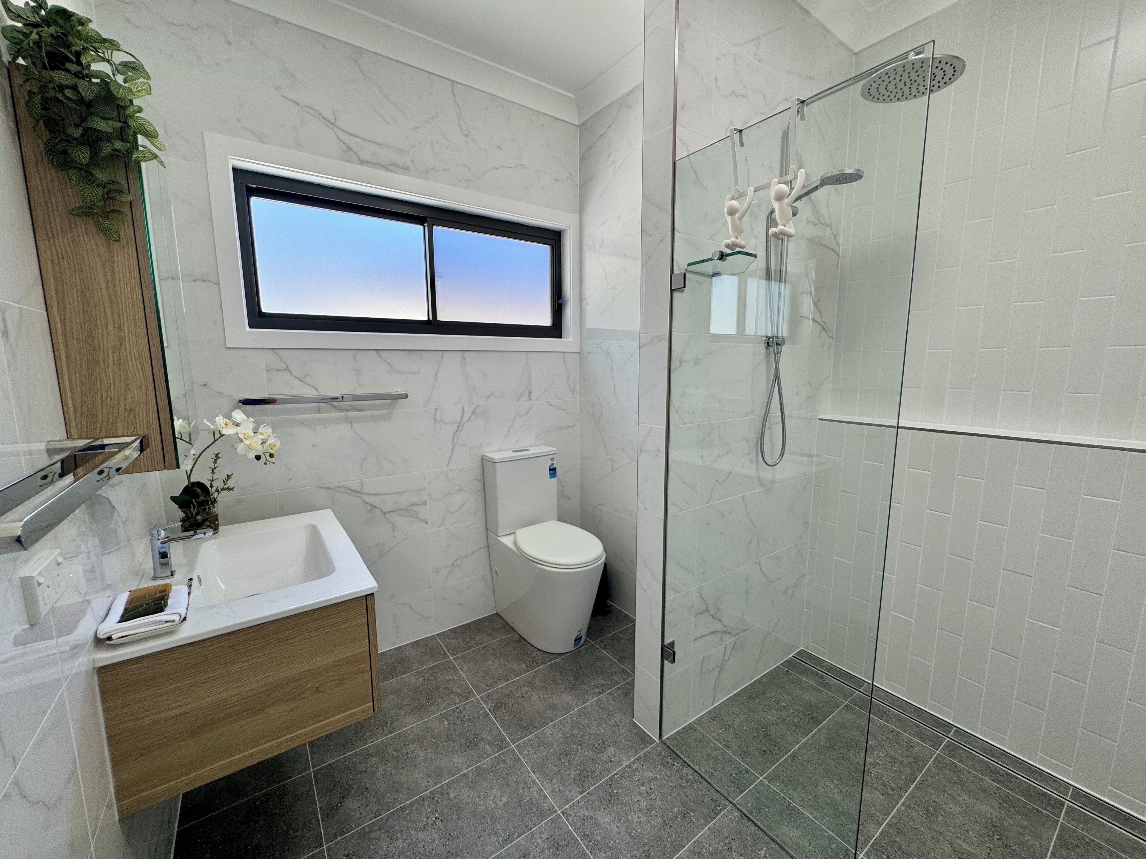 Een moderne badkamer met een marmeren wastafel, een toilet, een douche met glazen wand en een raam boven de wastafel.