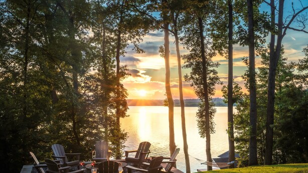 A lakeside deck with Adirondack chairs and a table, surrounded by trees and overlooking a body of water. - Lakefront Suite - Spa, Firepit, Kayaks & PBall (Macon)