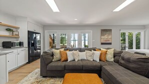 A modern living room with a sectional sofa, a kitchen with white cabinetry, and a large window offering a view of the outdoors. - Lakefront Suite - Spa, Firepit, Kayaks & PBall (Macon)