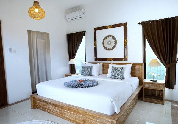 A neatly arranged hotel room with a large bed, a bedside table with a lamp, a wall-mounted air conditioner, and a decorative mirror above the bed.