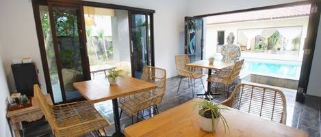 A dining area with a wooden table and wicker chairs, a hanging light fixture, and a view of an outdoor swimming pool.