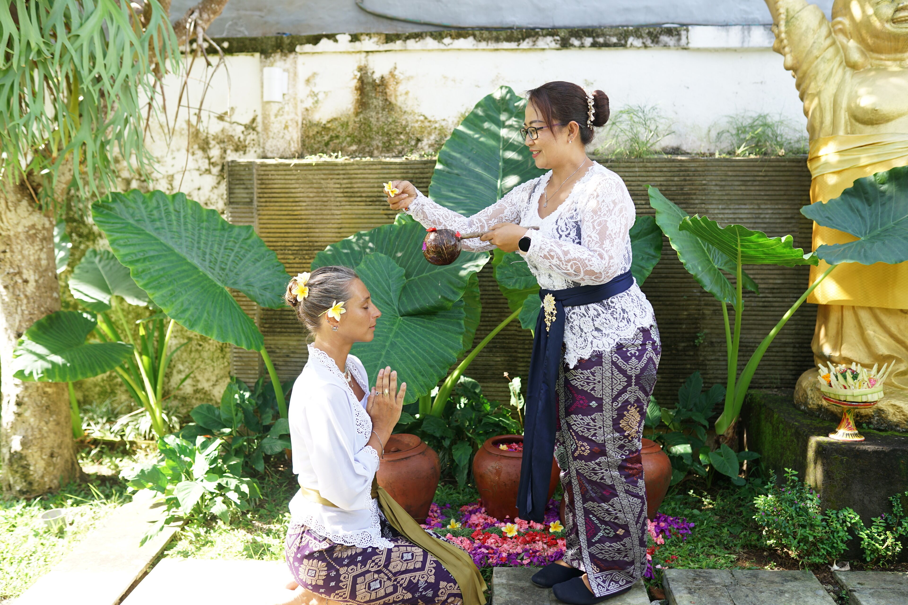 Two women in traditional attire in a garden with large plants and a golden statue.