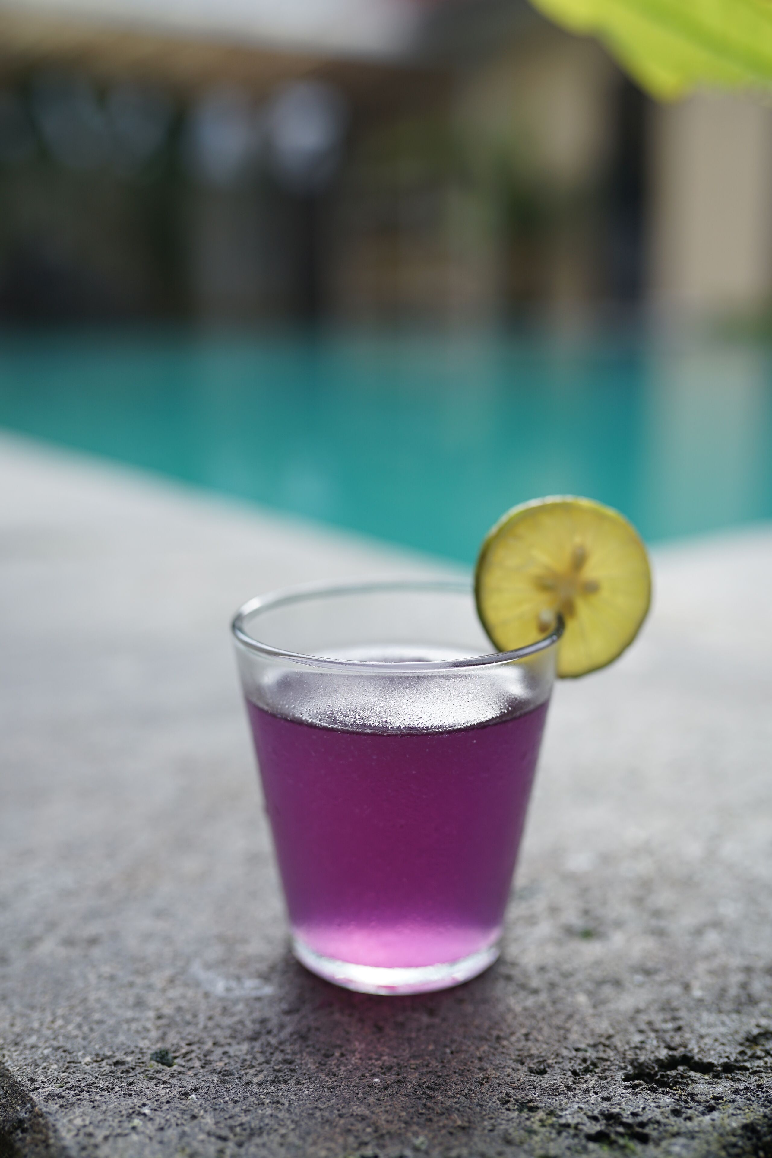 Ein Glas lila Getränk mit Limettenscheibe auf einer Steinoberfläche in der Nähe eines Pools.