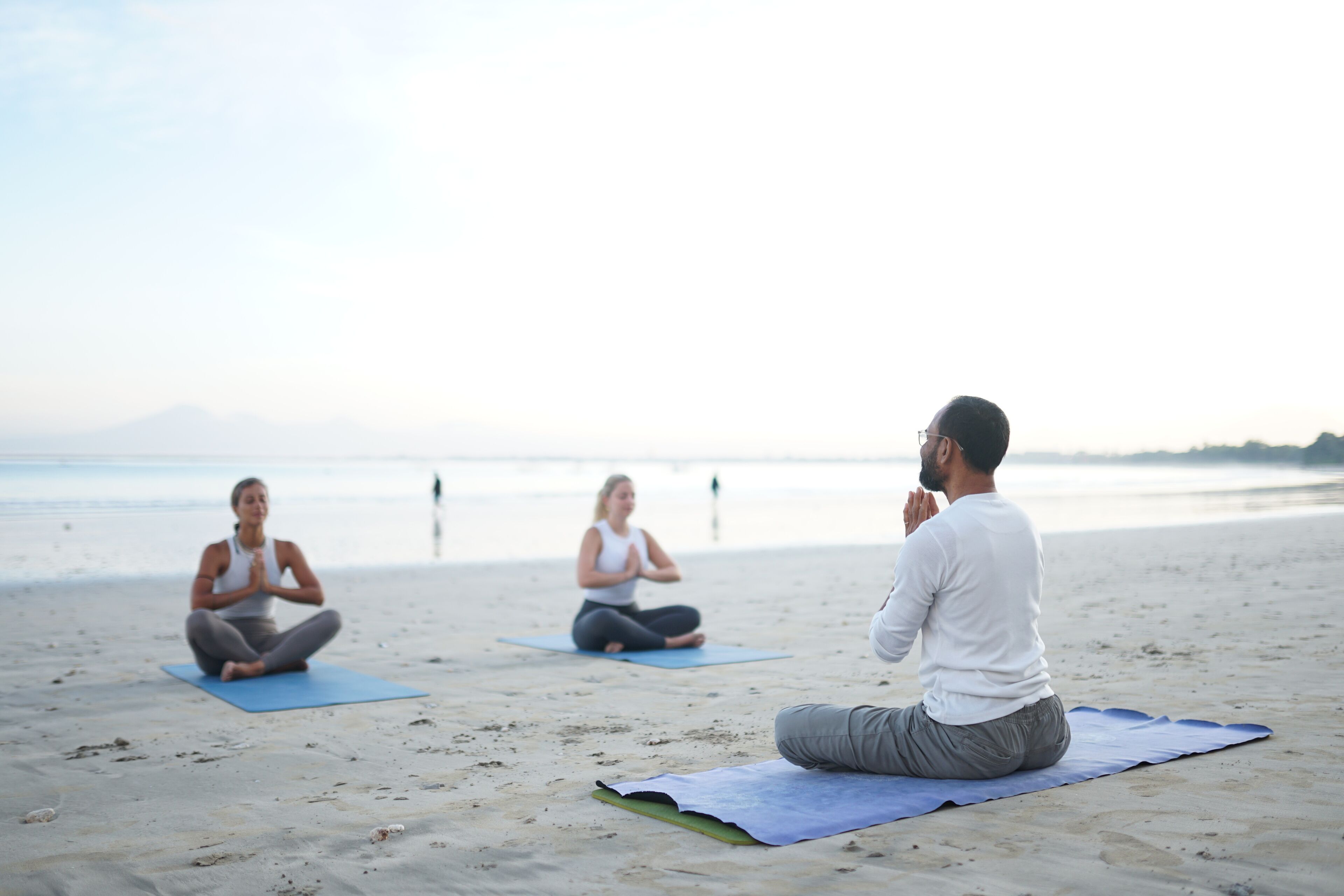Three individuals meditating on yoga mats on a sandy beach with a calm sea and mountains in the background.