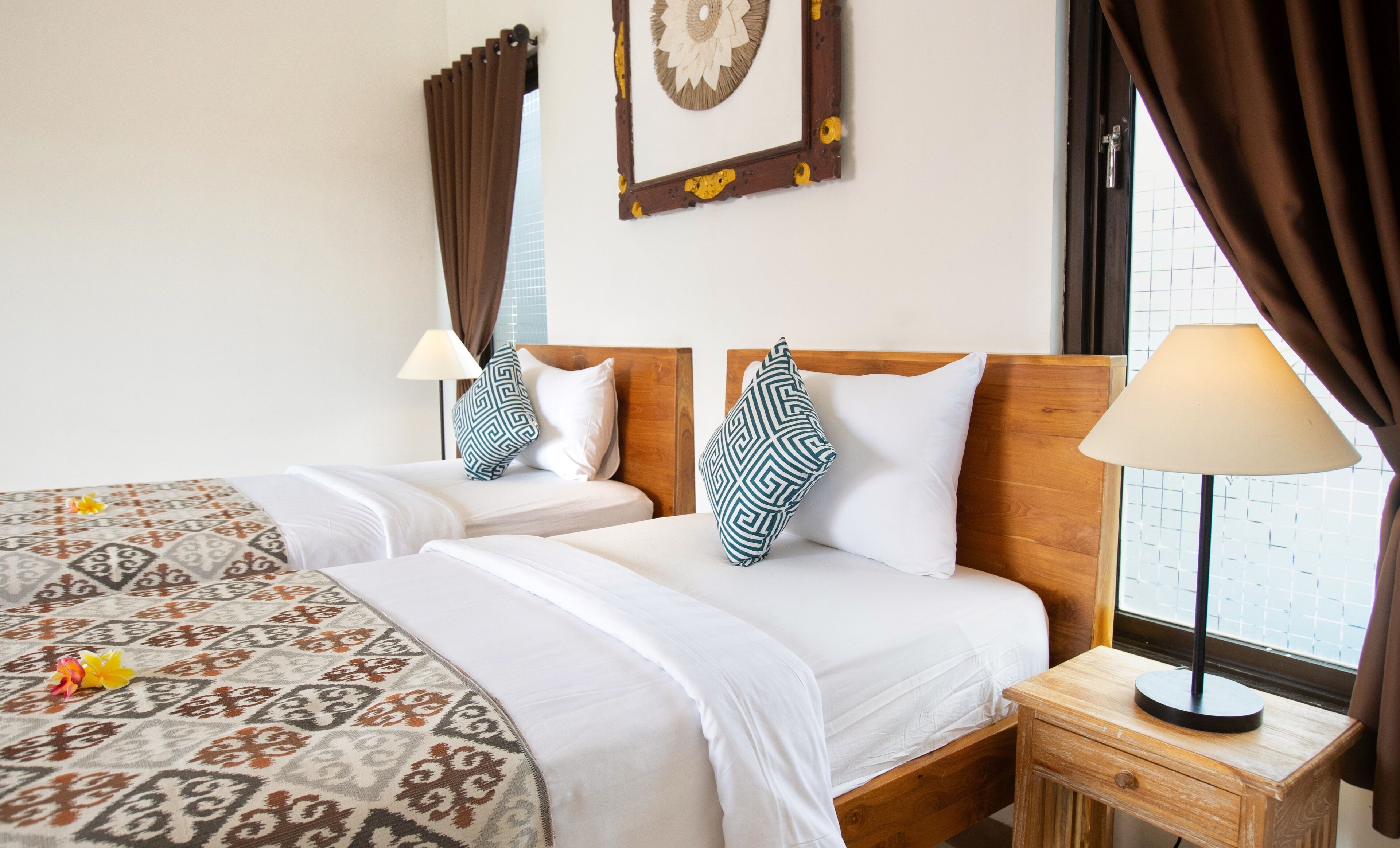 A hotel room with two beds, patterned bedspreads, wooden headboards, a nightstand with a lamp, and a window with curtains.