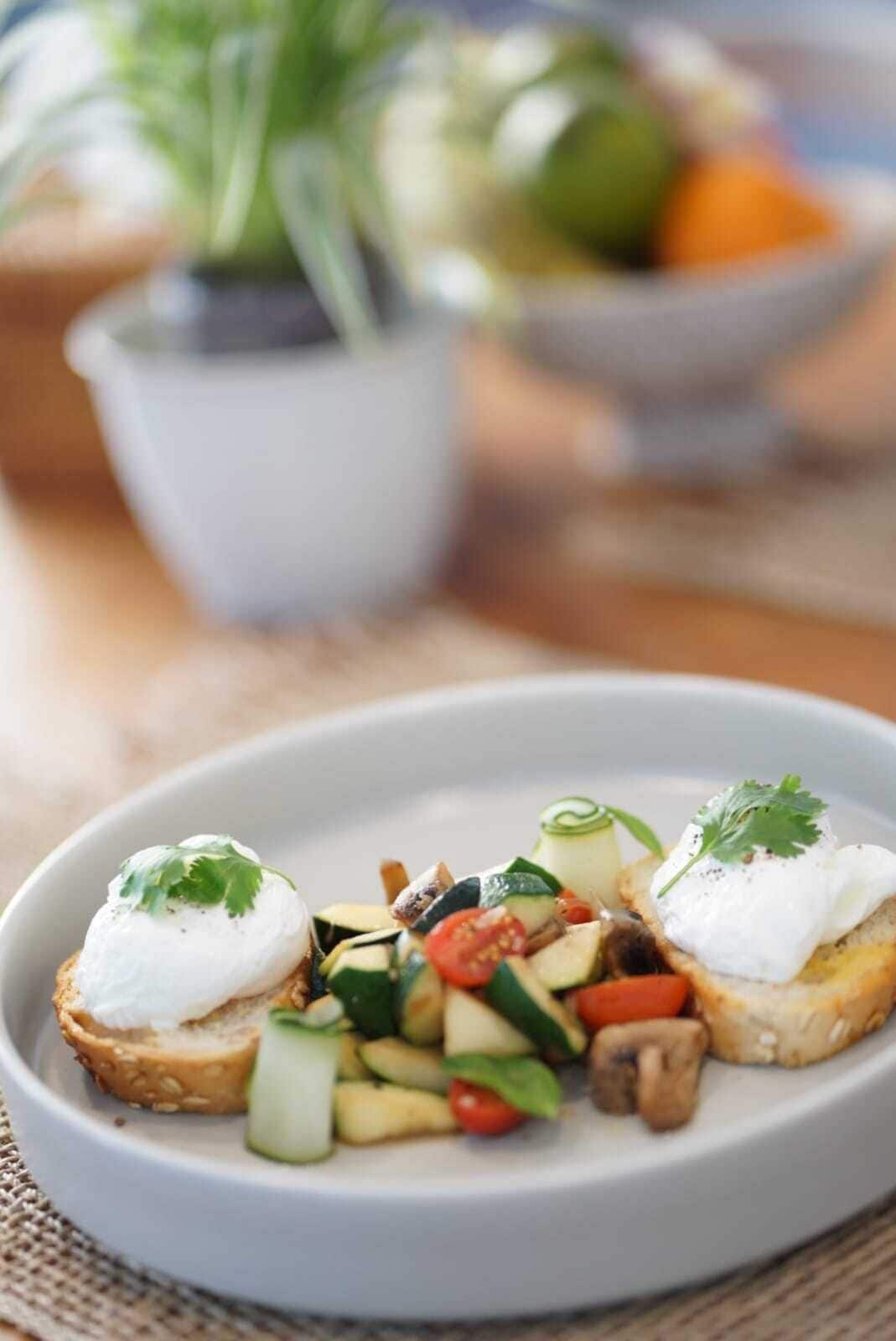 A plate with a vegetable and egg salad on toast, garnished with parsley, with a blurred background including a plant and a bowl.