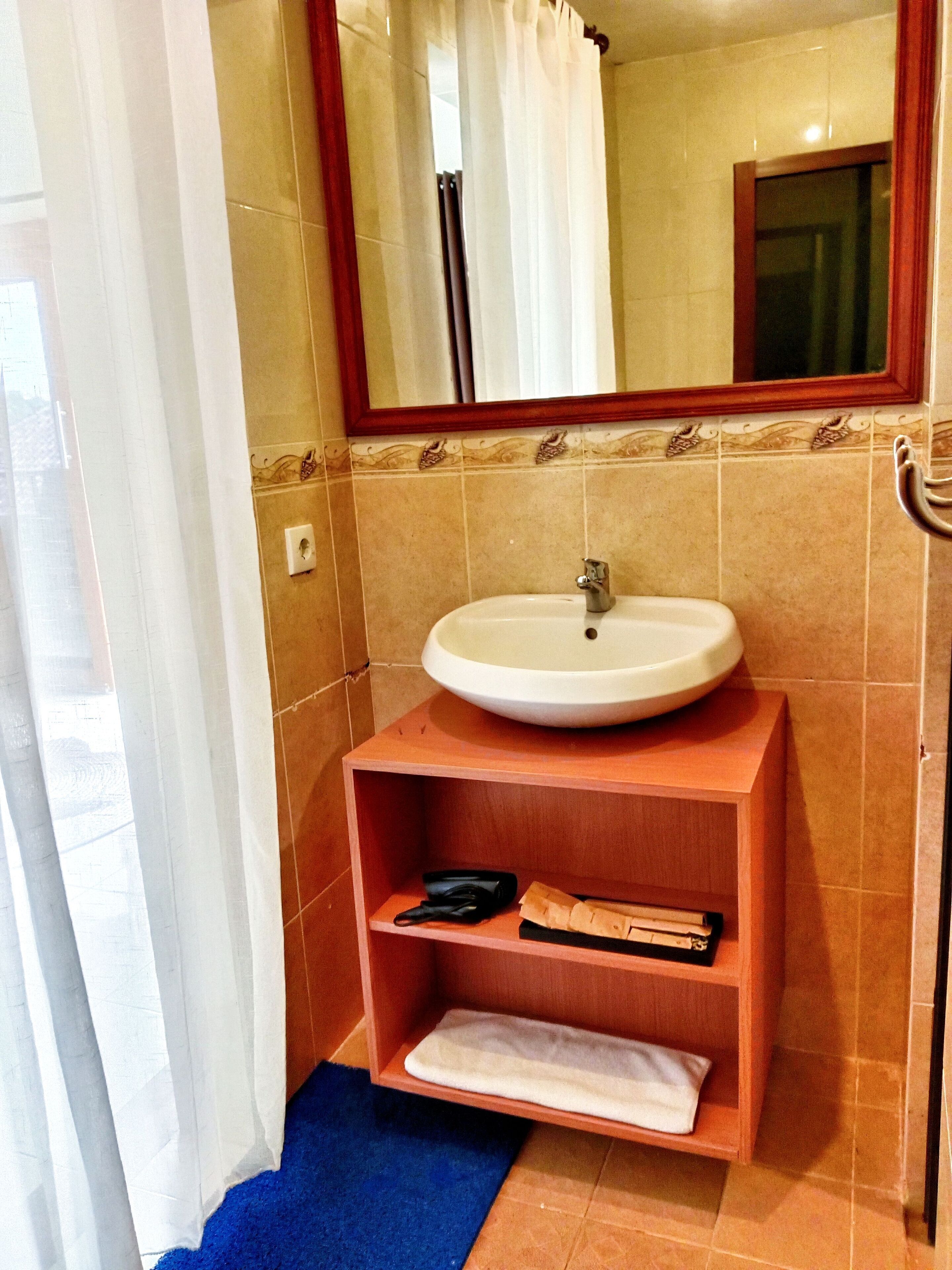 A bathroom with a white sink, a mirror, and a wooden shelf with a towel and a book.