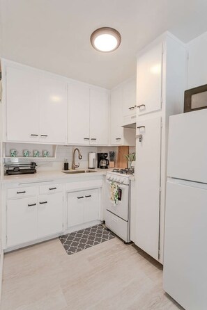 A modern kitchen with white cabinetry, a gas stove, a refrigerator, and a patterned rug. - King Suite - Walk to Beach - Patio + Parking (Oceanside)