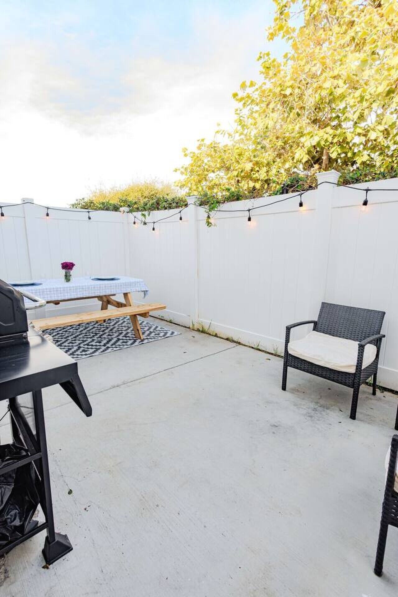 Een terras met een witte hek, een tafel met een blauwe tafelkleed, een stoel en een barbecue.