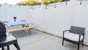 Een terras met een witte hek, een tafel met een blauwe tafelkleed, een stoel en een barbecue.