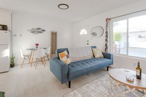 Un salon avec un canapé bleu, un coin repas avec une table et des chaises blanches, et une cuisine équipée d’un réfrigérateur.