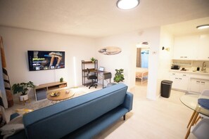 Una sala de estar con un sofá azul, un televisor montado en la pared, un escritorio pequeño con una silla, y una zona de cocina con armarios blancos y un lavabo.