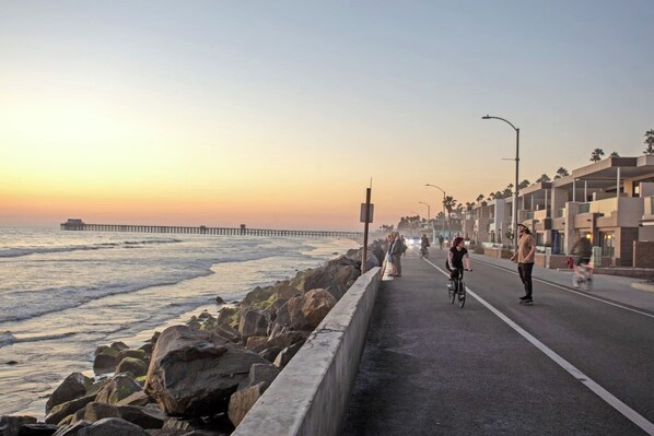 A coastal road with a bike lane, a pedestrian path, and a pier in the distance.