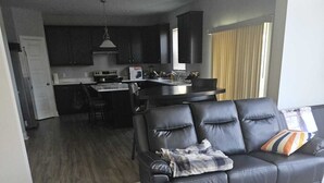 A modern living room with a black leather sofa, a dining area with a kitchen in the background, and a large window with blinds.