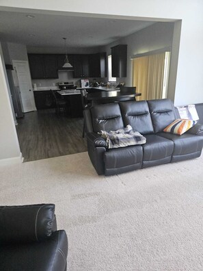 A modern living room with a black leather sofa, a dining area with a kitchen in the background, and a large window with blinds. - Spacious 5-bedroom house in delightful North Raleigh with AC, WiFi. (Raleigh)