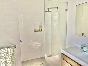 Une salle de bain moderne avec des murs recouverts de carreaux blancs, une douche en verre, un meuble-lavabo avec un miroir et une petite étagère contenant des articles de toilette.