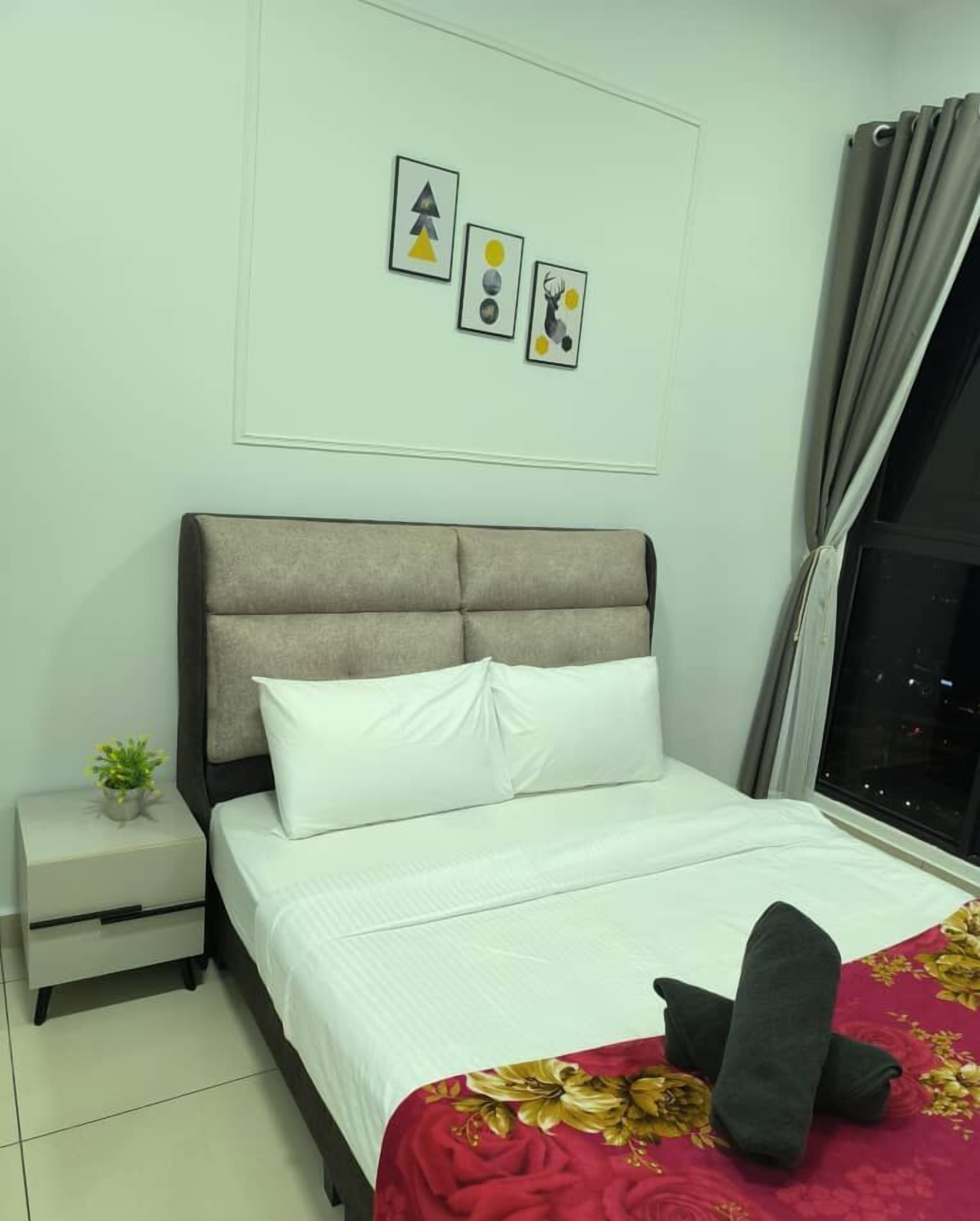 A neatly made bed with white bedding, a red floral bedspread, and a grey pillow. There is a small bedside table with a plant and a drawer. The room has a grey headboard and a window with grey curtains.