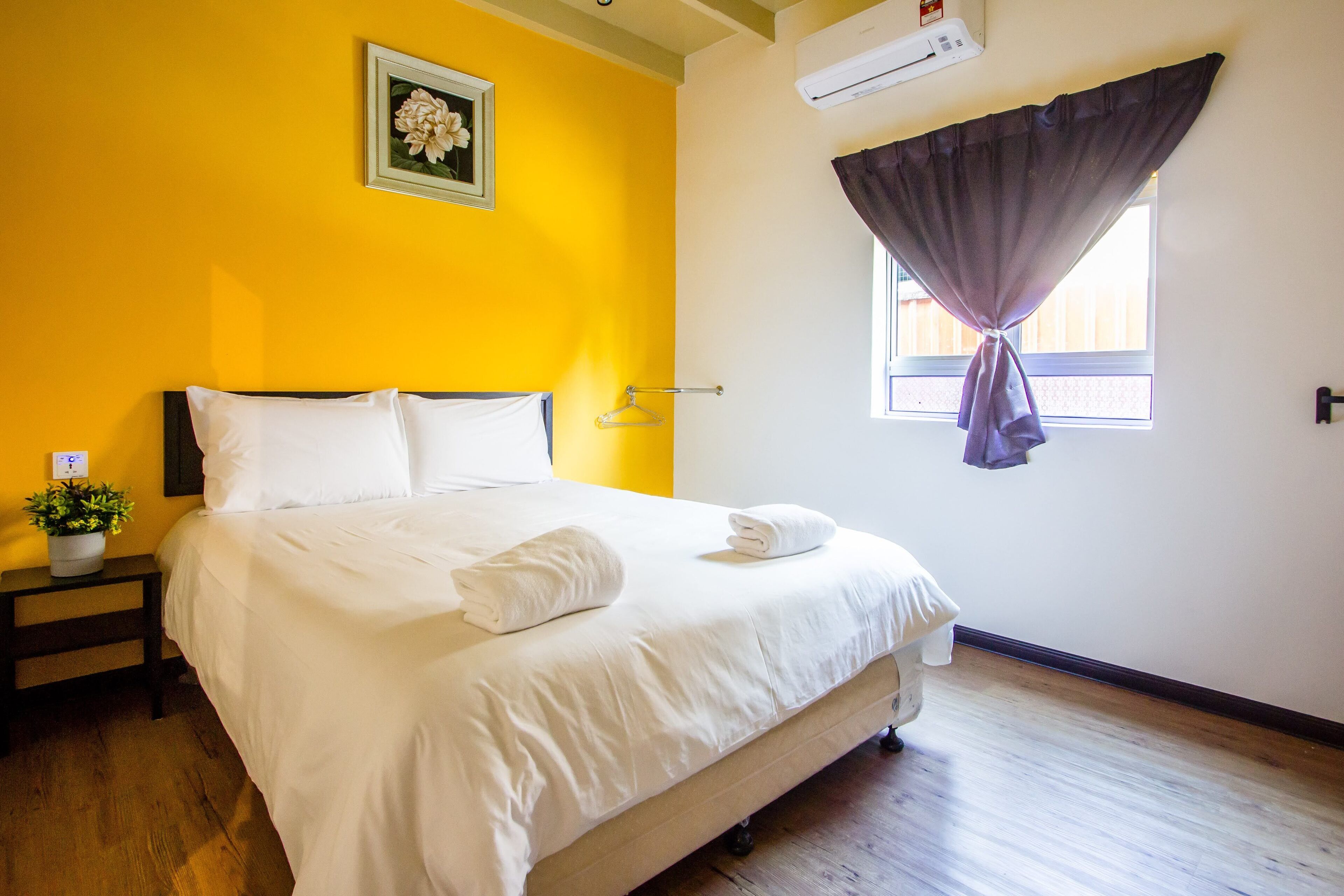 A neatly made bed with white linens and towels, a bedside plant, and a framed picture on the wall.
