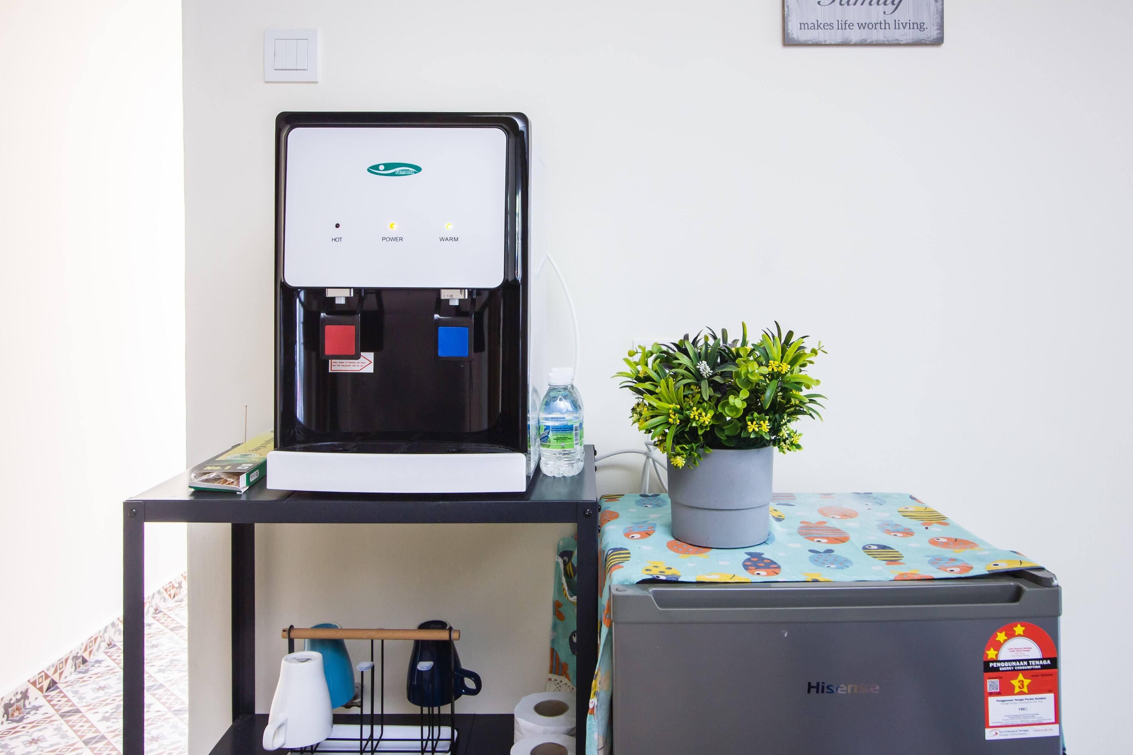 Un dispensador de agua, un refrigerador pequeño, una planta en maceta y una mesa con mantel colorido.