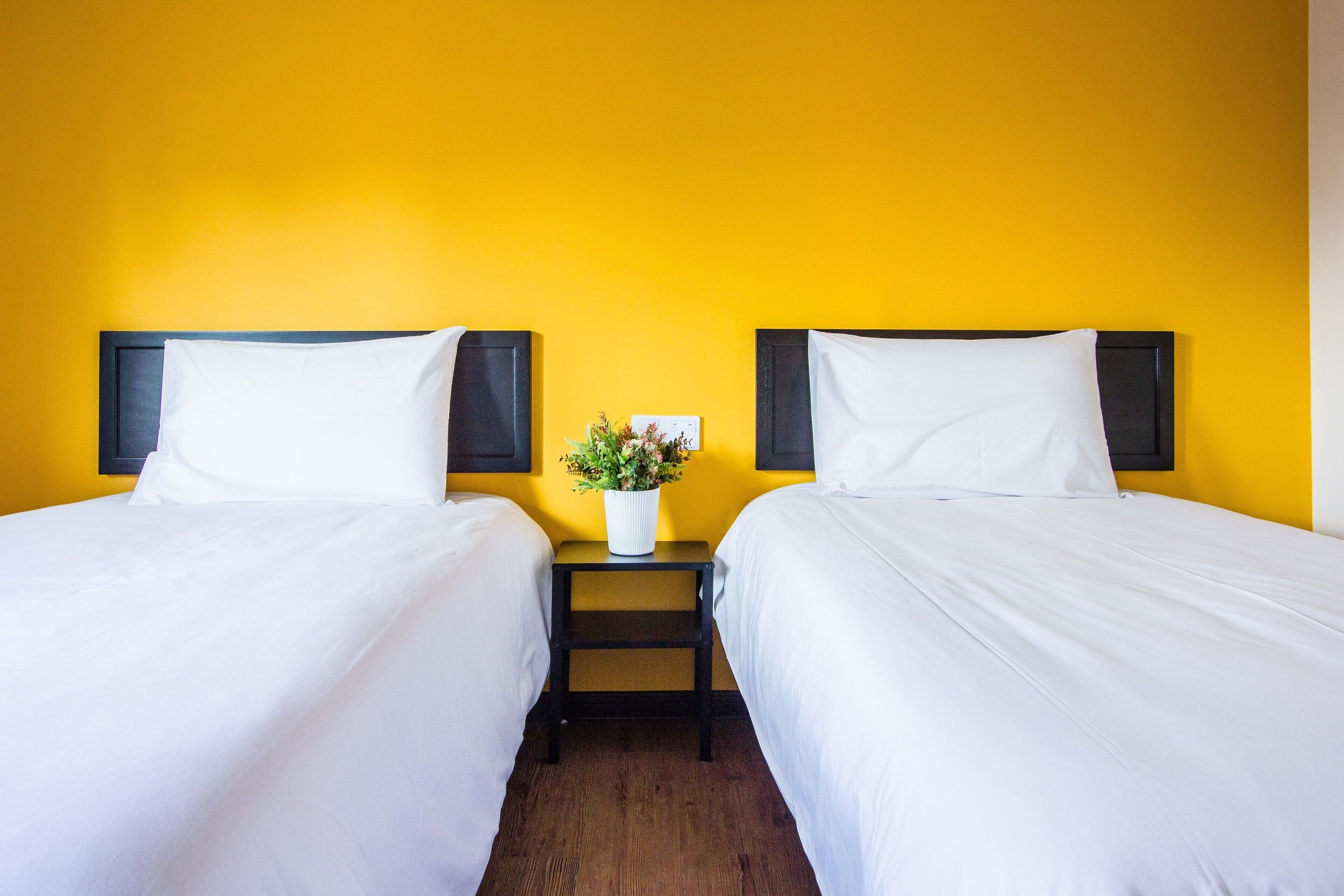 Habitación de hotel con dos camas, una mesita de noche con un jarrón de flores y una pared amarilla.