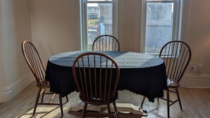 A dining area with a round table and four wooden chairs, large windows with a view of buildings, and a wooden floor.