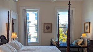 A bedroom with a four-poster bed, a ceiling fan, and large windows with a view of buildings outside.