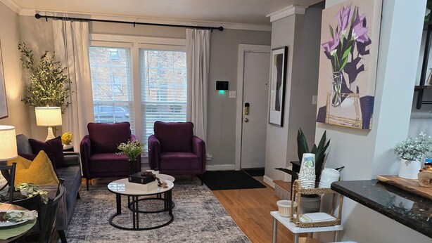A living room with a wooden floor, two purple sofas, a coffee table, a dining table with chairs, a white stool, and a small white table with decorative items. - A Pleasant Stay (Oak Park)