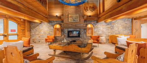 A log cabin interior with a stone fireplace, wooden furniture, and a chandelier.
