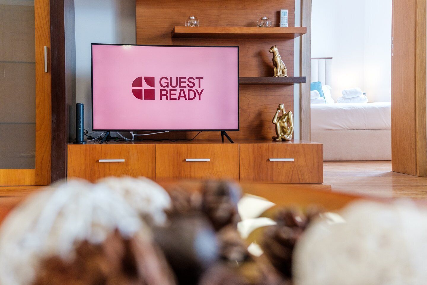 Ein modernes Wohnzimmer mit einem Flachbildfernseher, der den Schriftzug "GUEST READY" anzeigt, Holzmöbeln und dekorativen Accessoires.