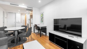 A modern living room with a round table, grey sofas, a flat-screen TV, and a kitchen area in the background. - Serain Residences Canberra Centre (Braddon)