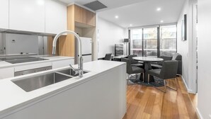 A modern kitchen with white cabinetry, a stainless steel sink, and a built-in oven. There is a dining area with a round table and chairs. - Serain Residences Canberra Centre (Braddon)