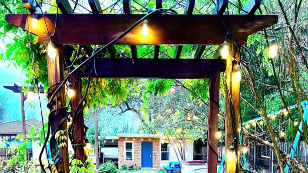 A stone pathway leading to a wooden pergola adorned with string lights, surrounded by a wooden fence and greenery.