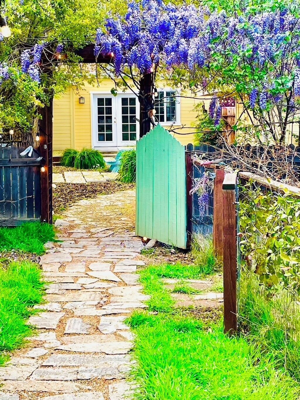 En stenlagd gång leder fram till ett hus med en grön grind och en pergola som är prydd med lila blommor.
