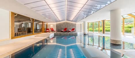 Una piscina interior moderna con paredes de vidrio transparente, sillas de descanso rojas y un techo espacioso con diseño de cuadrícula.