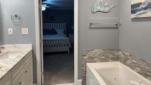 Une salle de bain avec une baignoire, une douche et des toilettes. Une porte donne sur une chambre à coucher équipée d’un lit et d’une table de chevet.