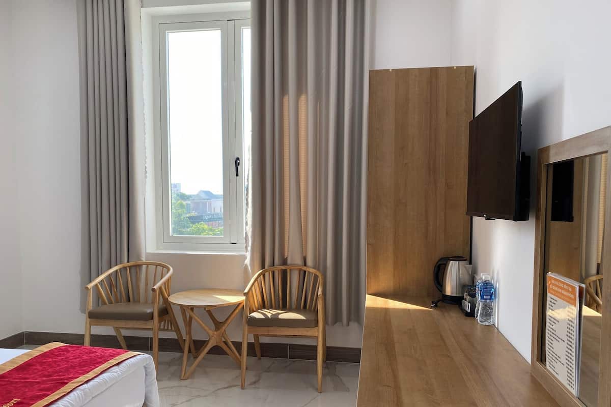 Une chambre d’hôtel avec un lit, une télévision, une table en bois, deux chaises, une fenêtre avec des rideaux et une vue sur l’extérieur.