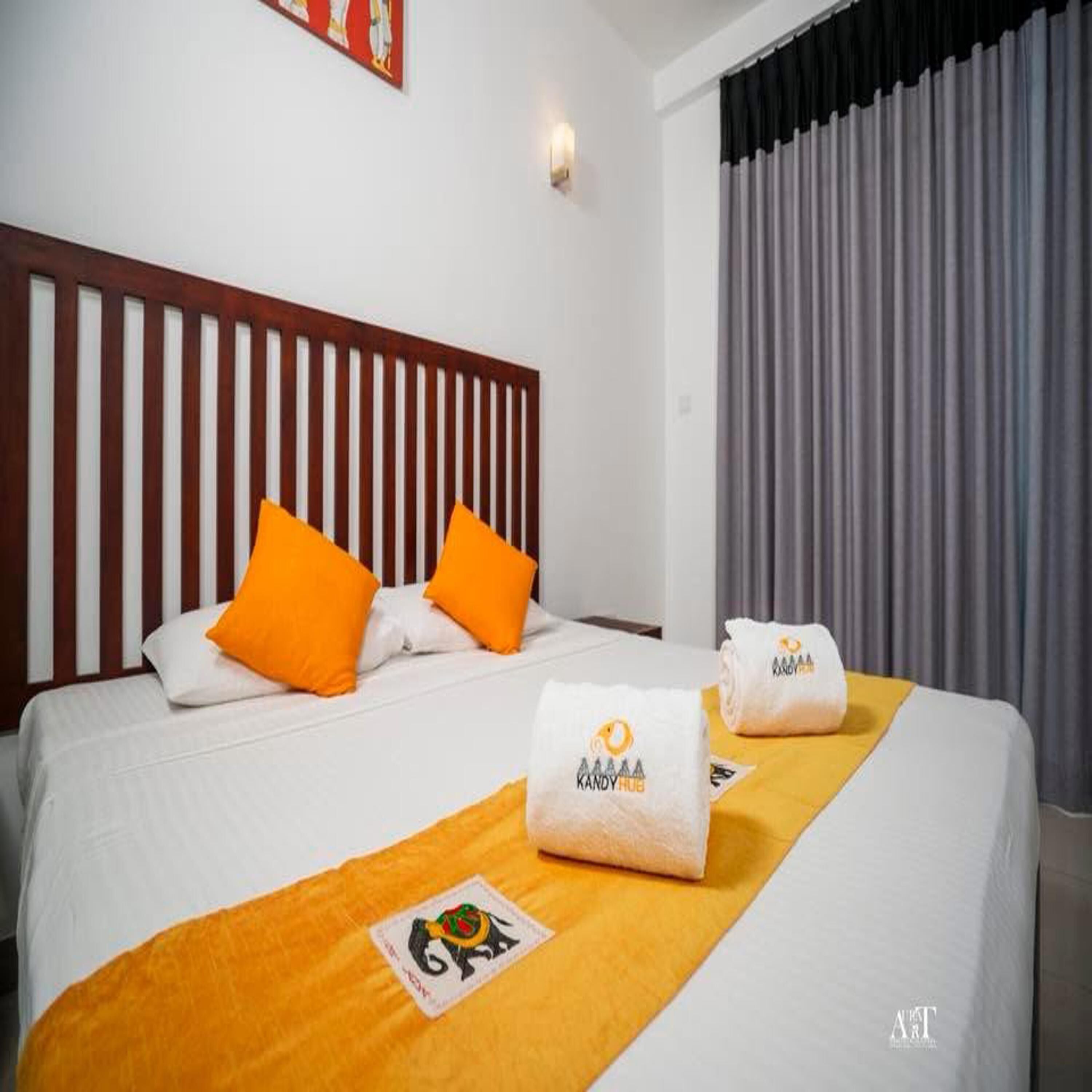 A hotel room with a bed, orange pillows, a headboard, a window with curtains, and a towel with branding.