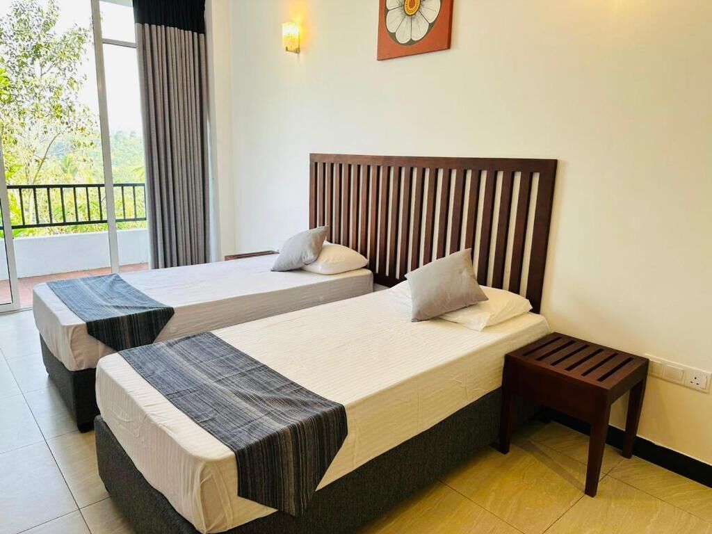 A hotel room with two beds, a small wooden nightstand, a window with curtains, and a wall-mounted light fixture.