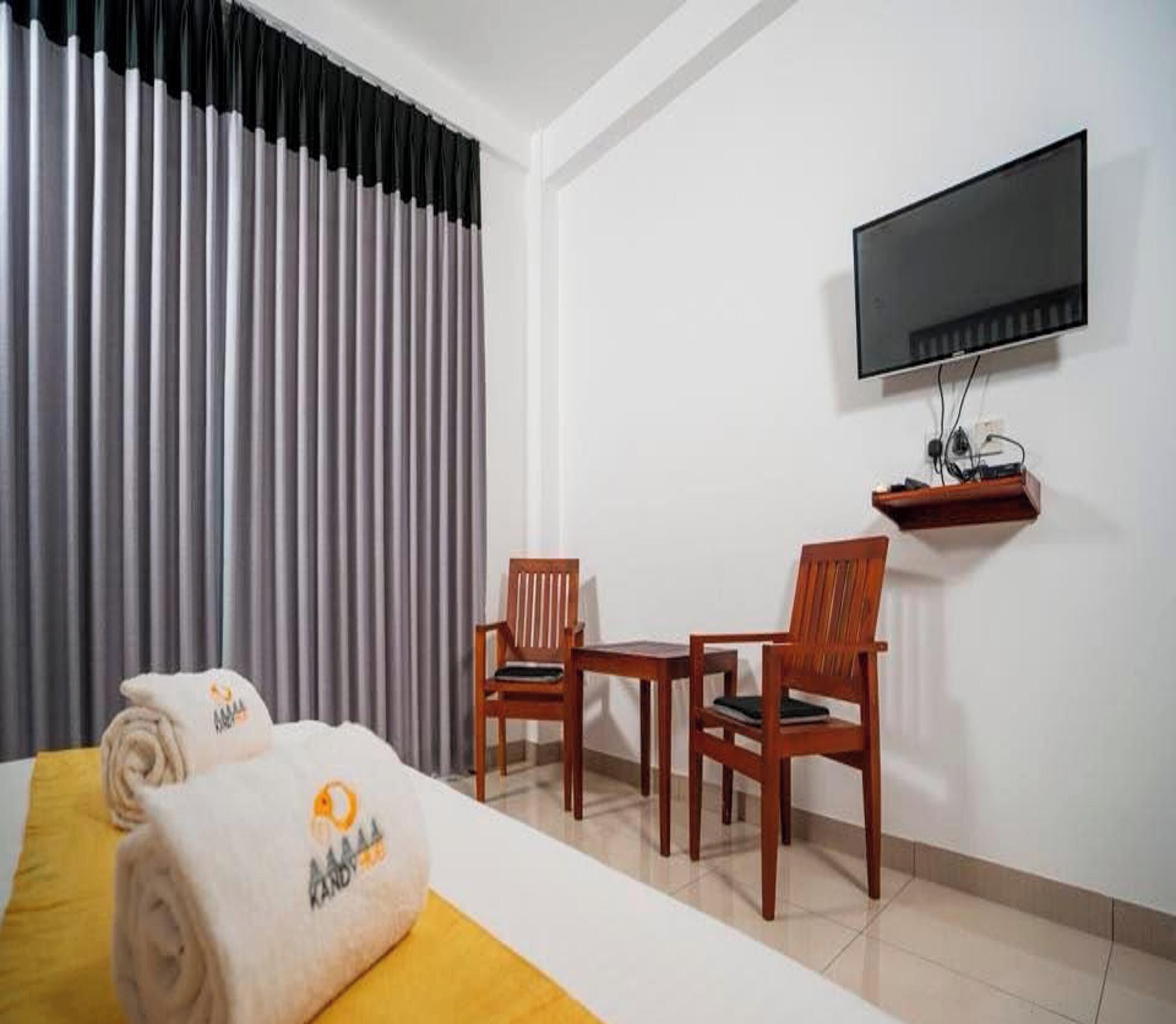 Quarto de hotel com uma cama, duas cadeiras de madeira, uma televisão presa na parede e uma janela com persianas verticais.