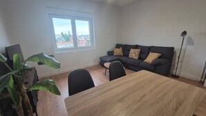 Ein modernes Wohnzimmer mit einem Holzesstisch, einem grauen Sofa mit gemusterten Kissen, einem großen Fenster und einer Topfpflanze.