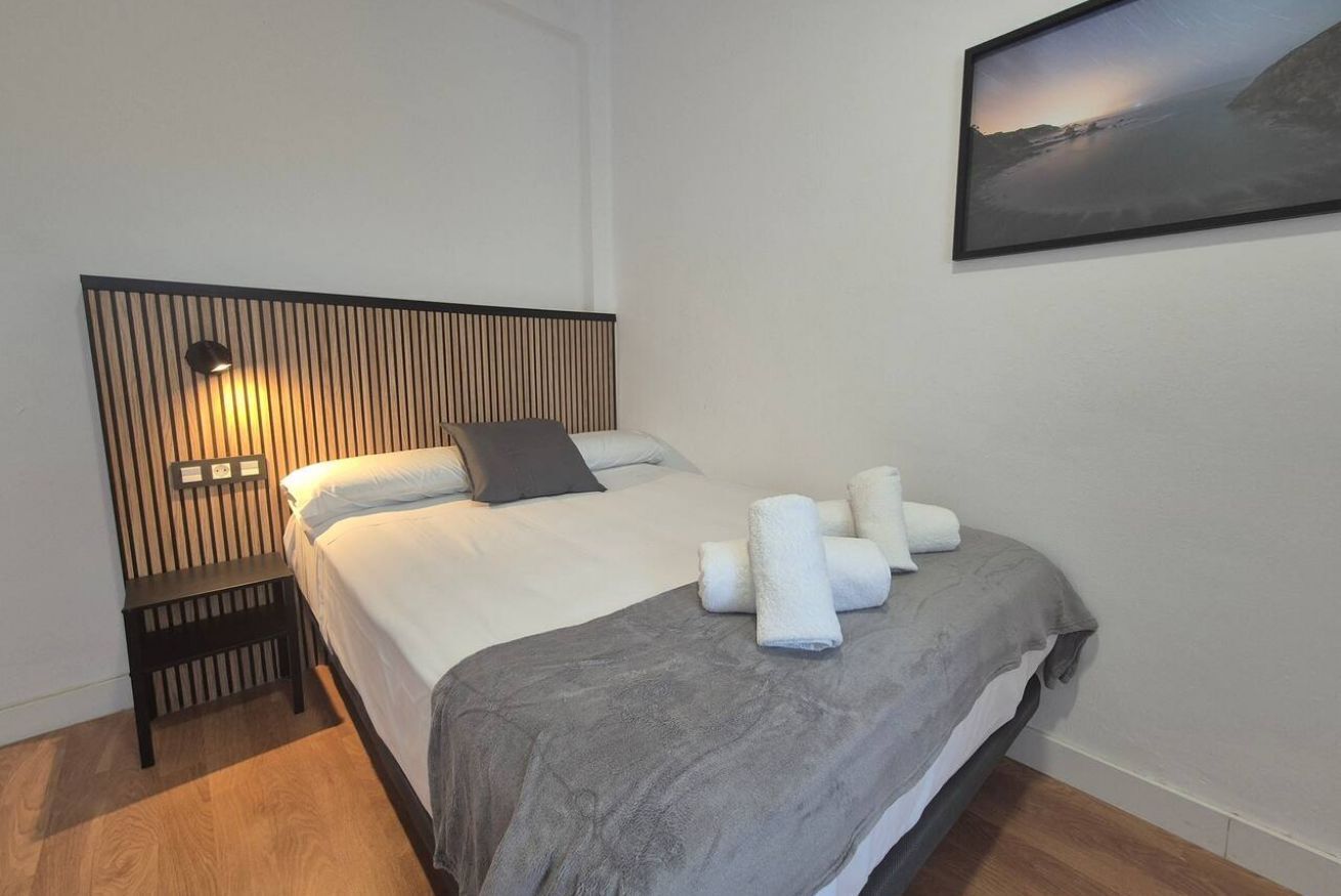Una cama bien hecha con una manta gris y toallas enrolladas, una lámpara de noche y un cuadro en la pared.