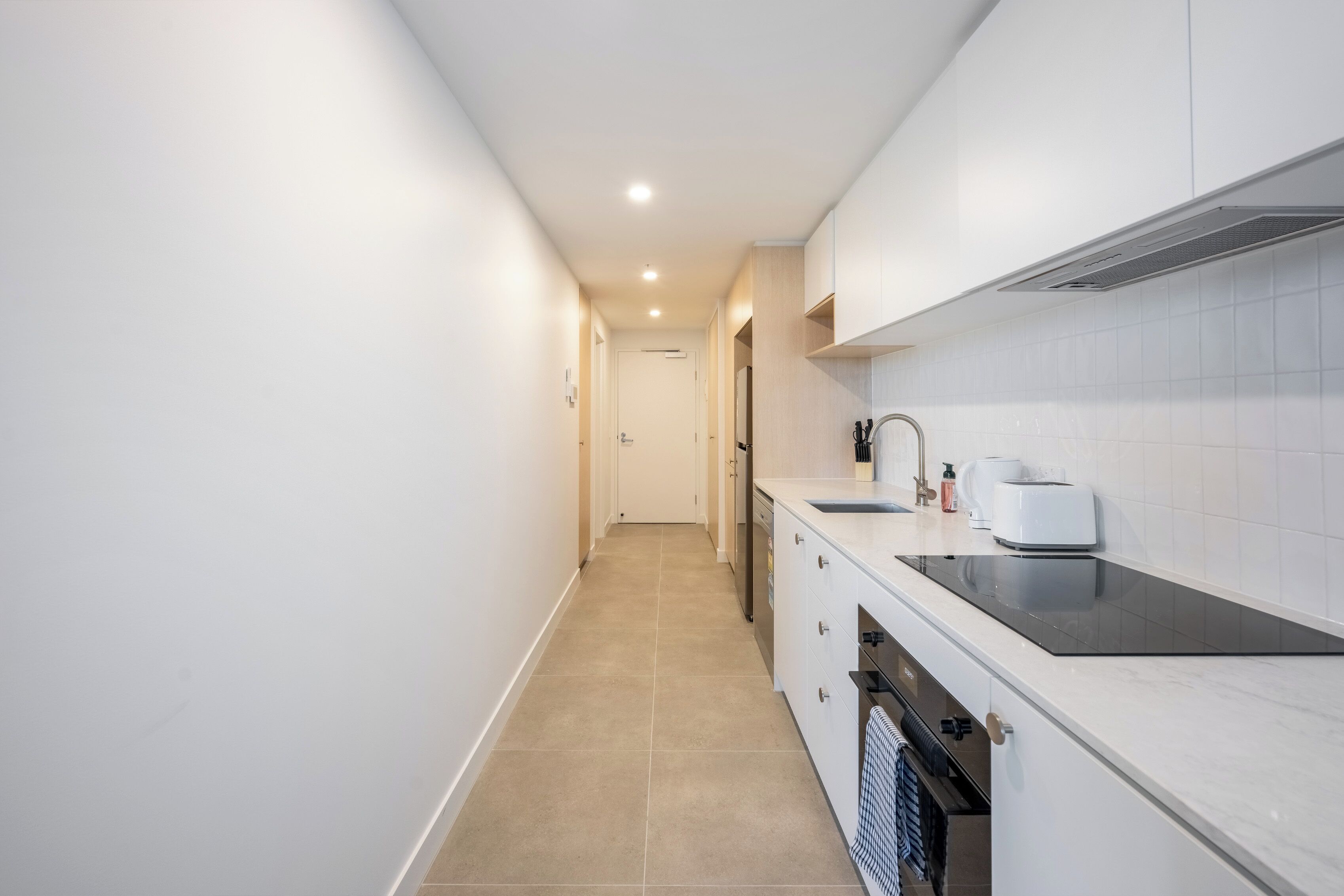A narrow, well-lit kitchen with white cabinets, a black countertop, and a tiled backsplash.