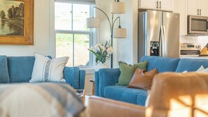 Un salon moderne avec un canapé bleu, une table basse et un vase rempli de fleurs. Une fenêtre offre une vue sur la verdure à l’extérieur.