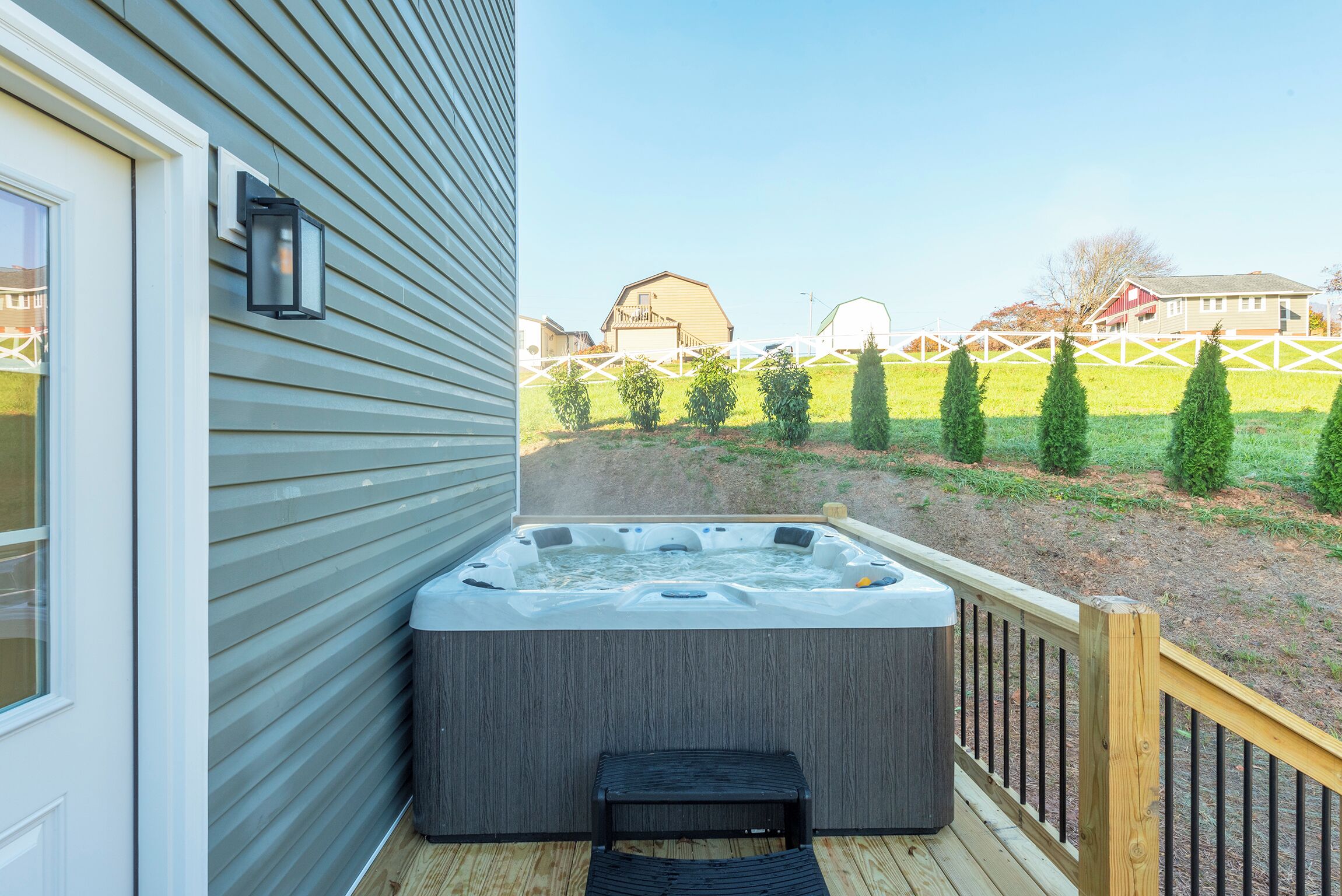 Un jacuzzi en una terraza de madera con vista a un jardín cercado y edificios lejanos.
