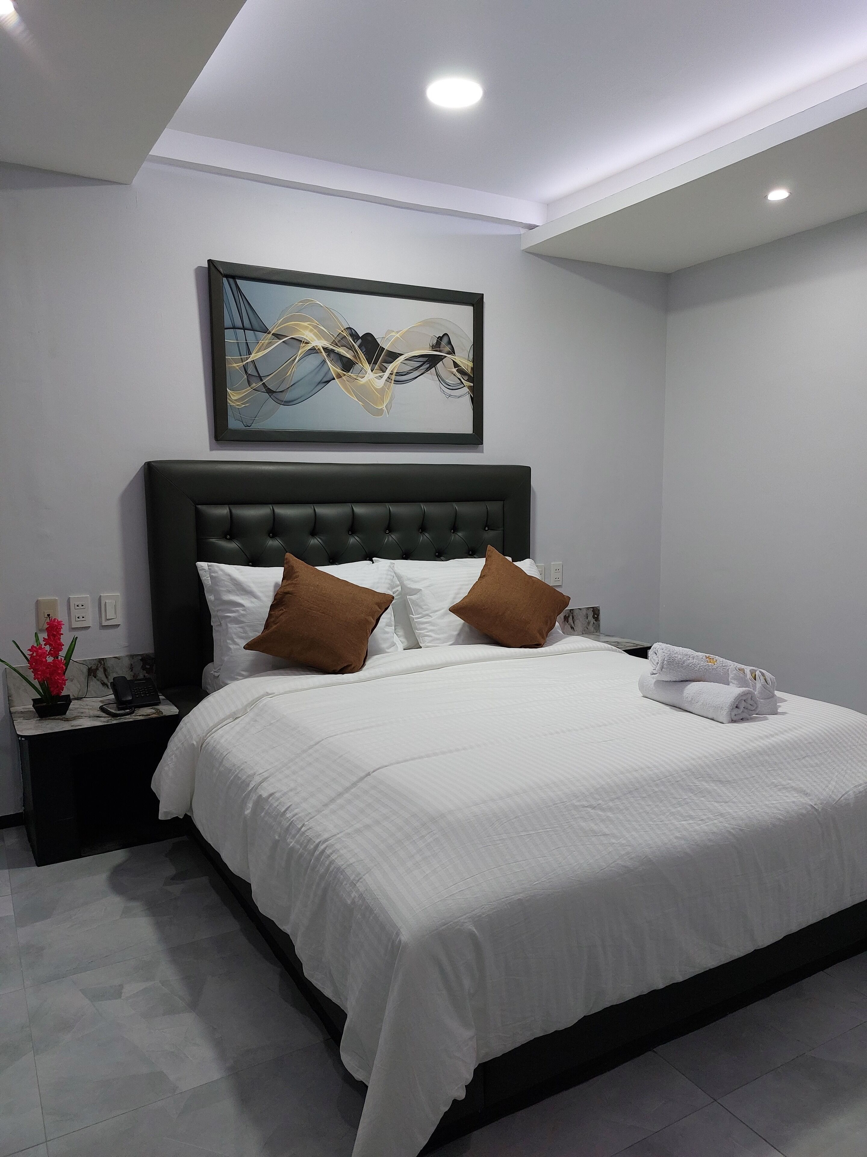 A modern bedroom with a large bed, a black headboard, white bedding, and a painting above the bed.